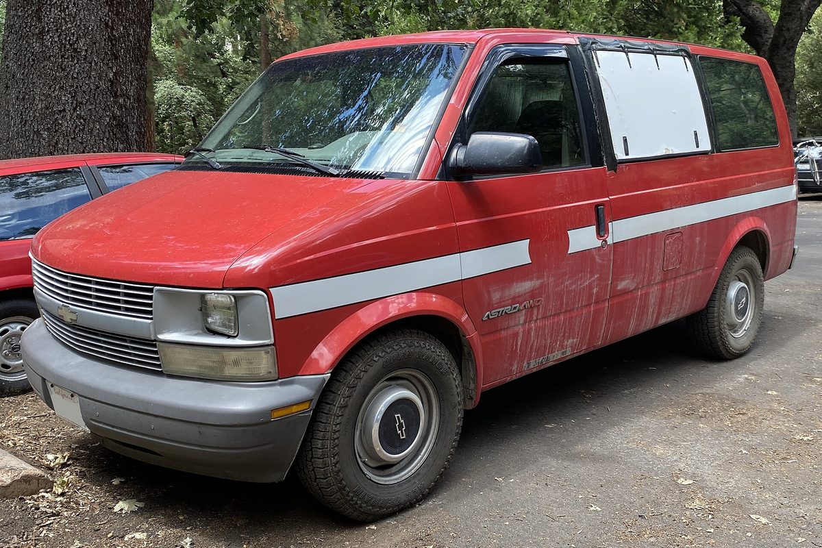 A faded red 1995 Chevrolet Astro AWD van with streaked, peeling paint on its doors, a white side stripe and a covered rear side window, parked on gravel and dirt under tall trees