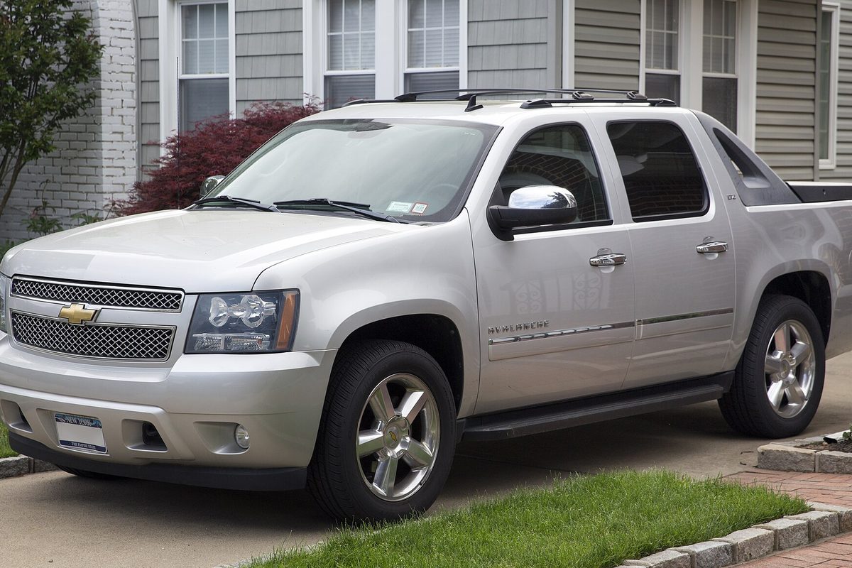 A silver 2011 Chevrolet Avalanche LTZ with chrome wheels and roof rails, parked in a driveway in front of a gray-sided house
