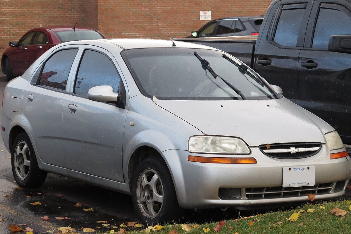 A silver 2005 Chevrolet Aveo LT sedan on wet pavement scattered with fallen leaves, parked beside a dark pickup truck