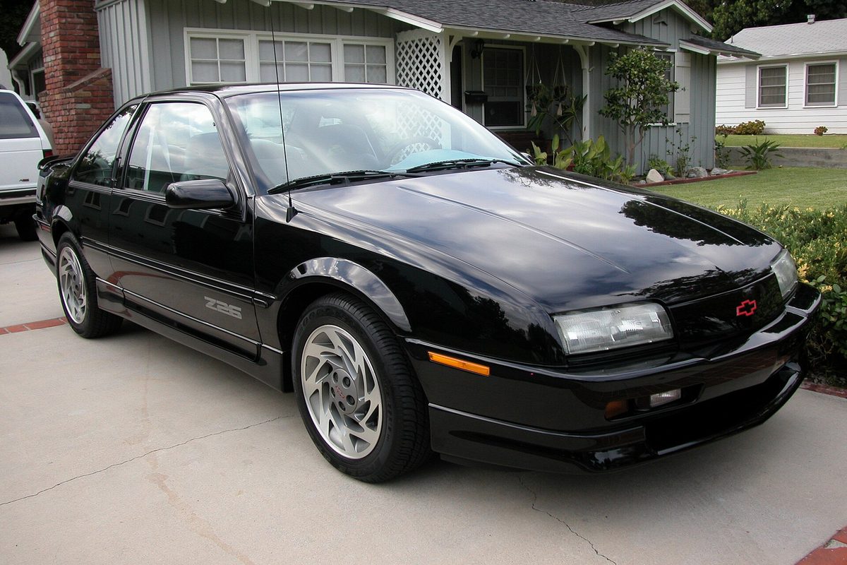 A black 1996 Chevrolet Beretta Z26 coupe with silver alloy wheels, parked in a concrete driveway beside a gray house