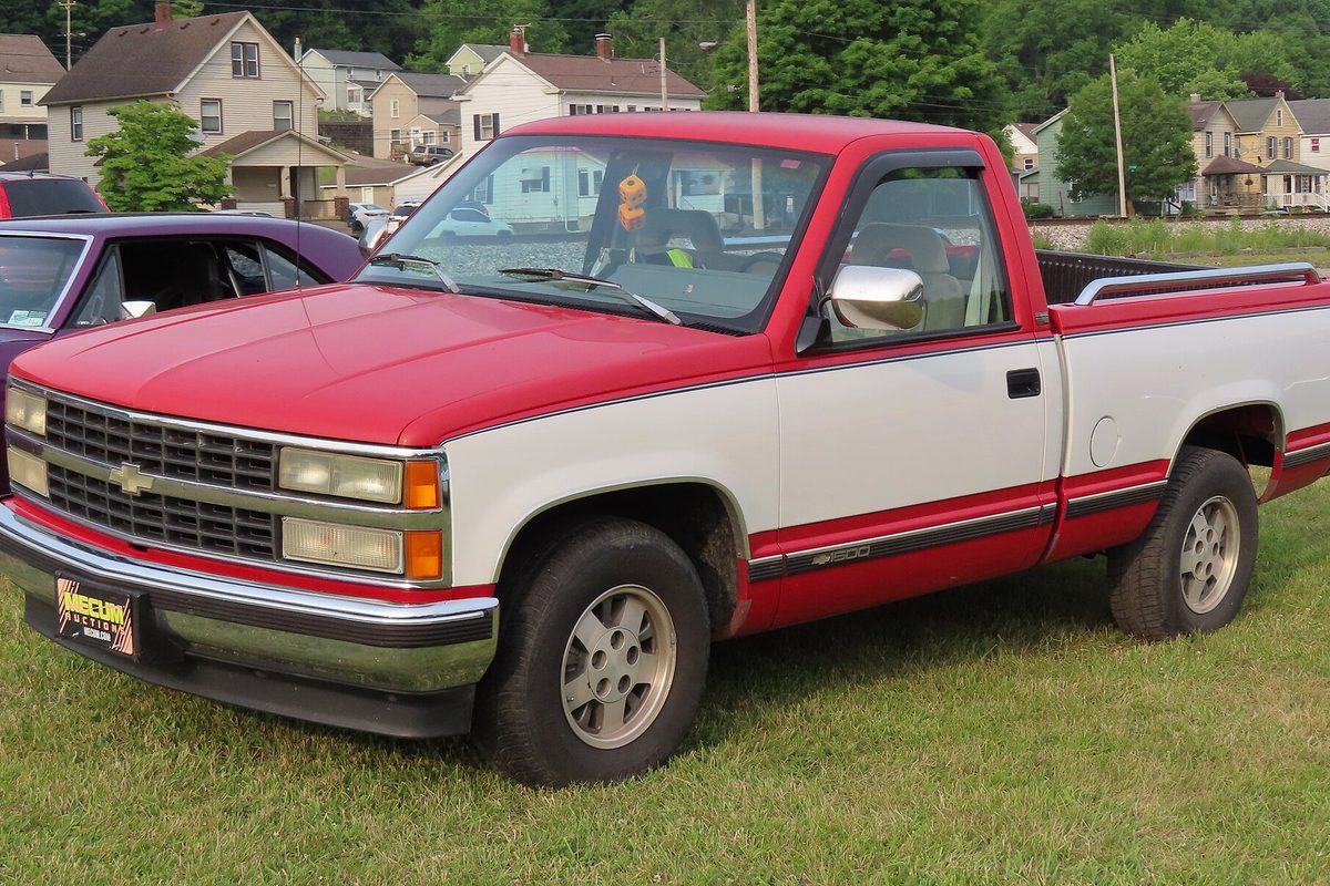 A red and white 1992 Chevrolet C1500 regular-cab pickup parked on the grass at a car show, with houses on the hill behind it