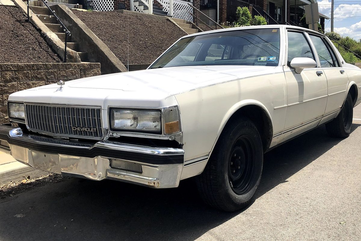 A white 1989 Chevrolet Caprice Classic sedan with a dented chrome front bumper and black steel wheels, parked at a curb below a hillside stairway