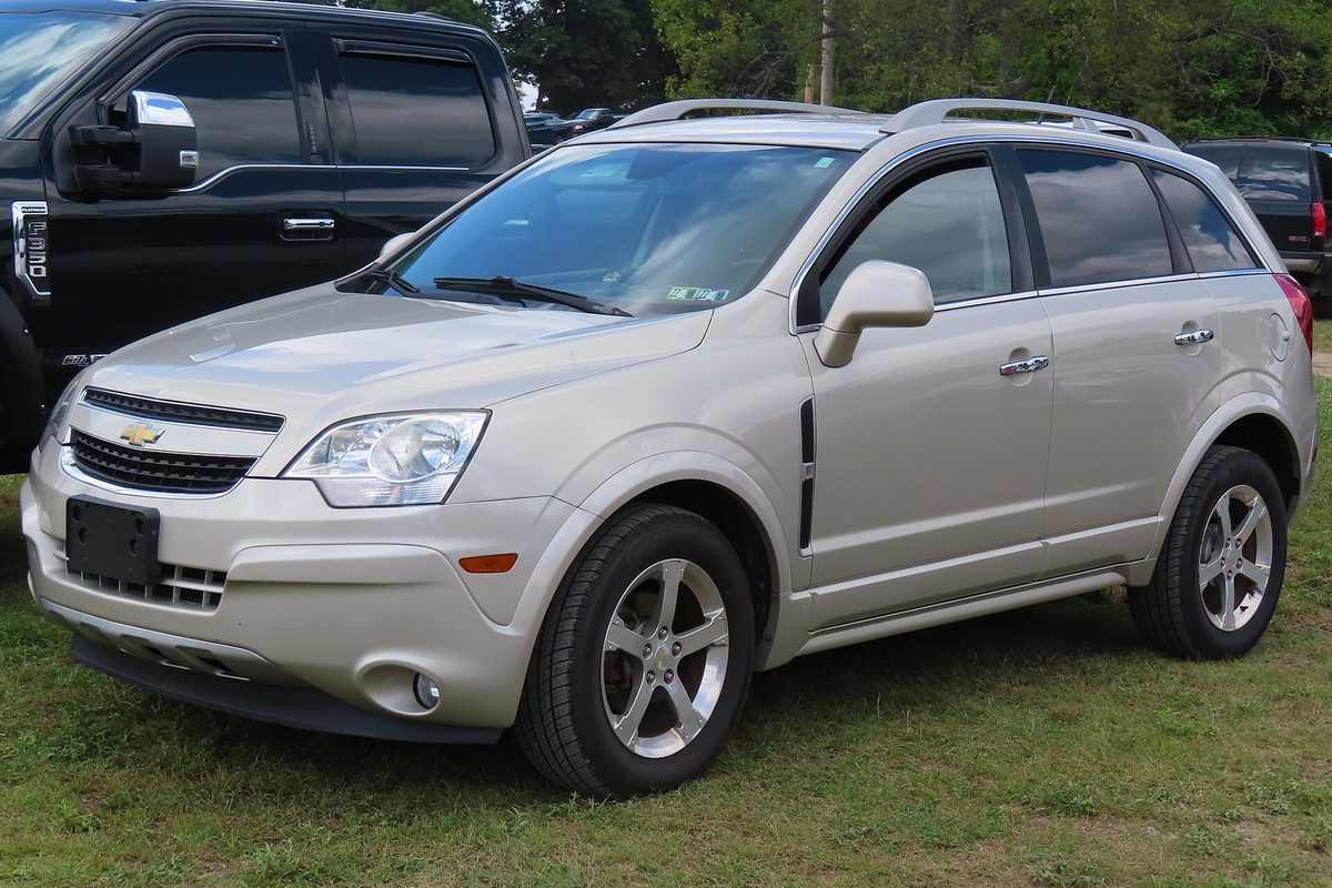 A champagne silver 2014 Chevrolet Captiva Sport LT parked on grass at an outdoor lot, next to a black pickup
