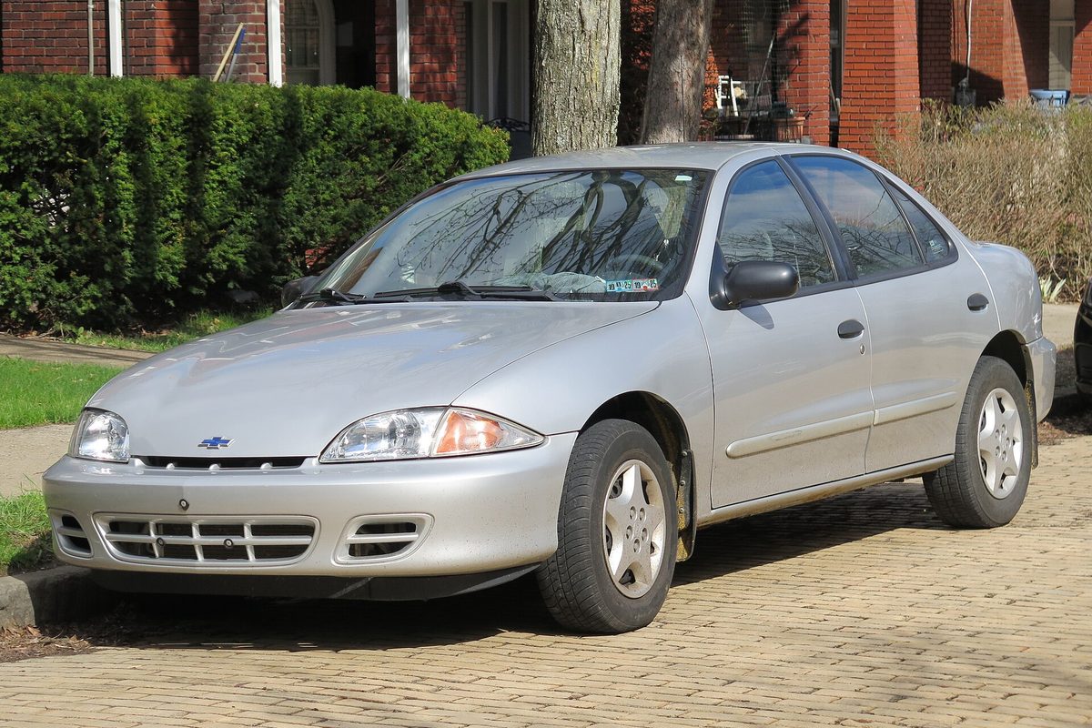 A silver 2000 Chevrolet Cavalier sedan with rust starting at the bottom of its rear door, parked on a brick street beside a hedge