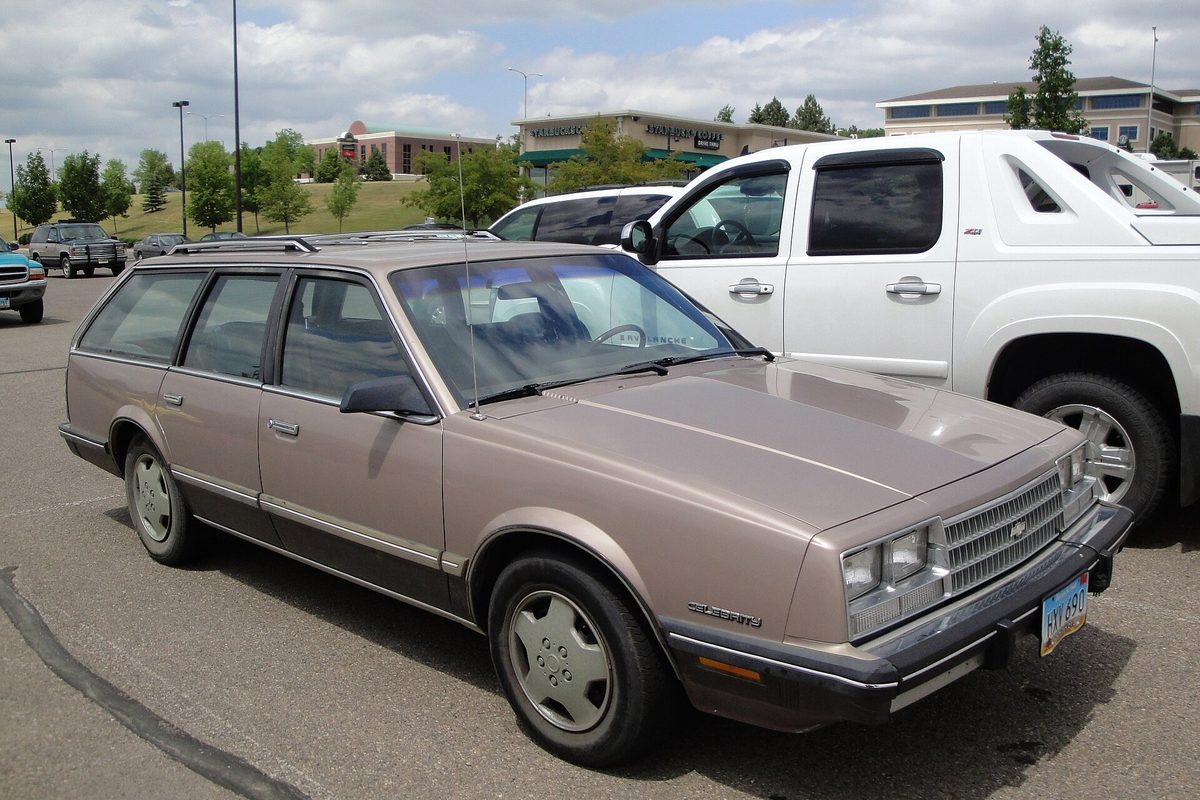 A tan 1985 Chevrolet Celebrity station wagon in a shopping center parking lot next to a white pickup