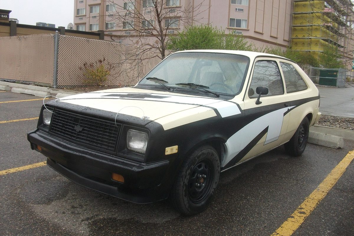 A 1986 Chevrolet Chevette hatchback in a hand-painted cream and black scheme with black steel wheels, parked in a wet lot near an apartment building