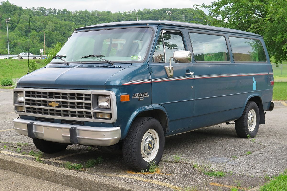 A teal 1994 Chevrolet Chevy Van 10 with side windows and a red pinstripe, parked in a cracked lot with weeds along the curb