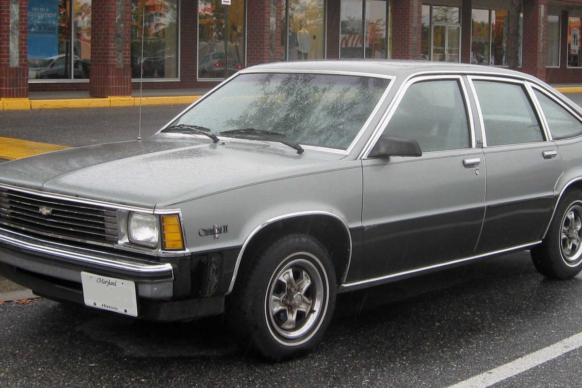A two-tone gray Chevrolet Citation II hatchback parked on a wet street in front of a row of shop windows