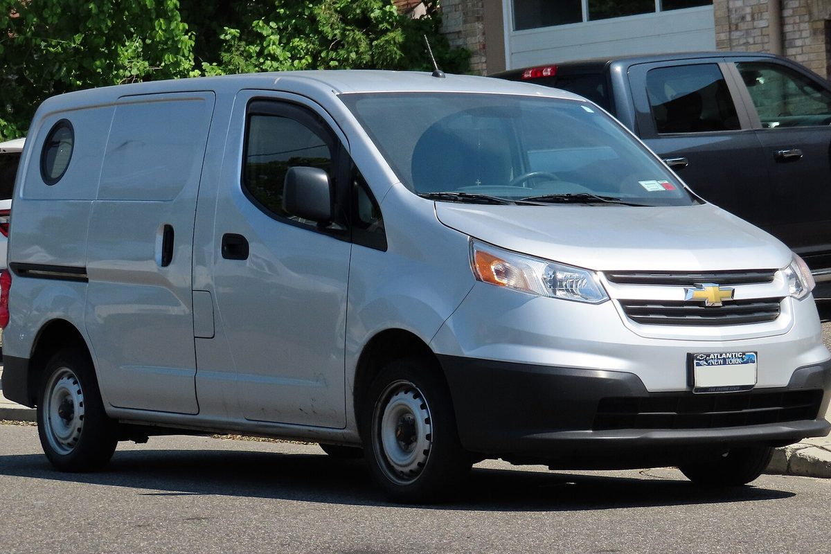 A silver 2015 Chevrolet City Express small cargo van with steel wheels and a round porthole window, parked at a curb under green trees