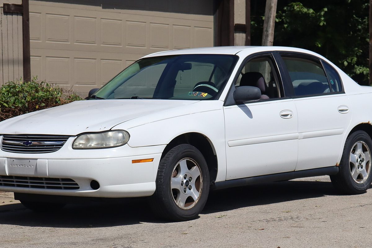 A white 2005 Chevrolet Classic sedan with rust bubbling at the rear wheel arch, parked in front of a closed garage door