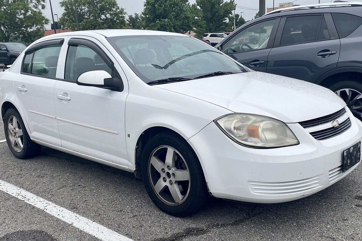 A white 2009 Chevrolet Cobalt sedan with worn five-spoke alloy wheels, parked in a lot beside a gray SUV