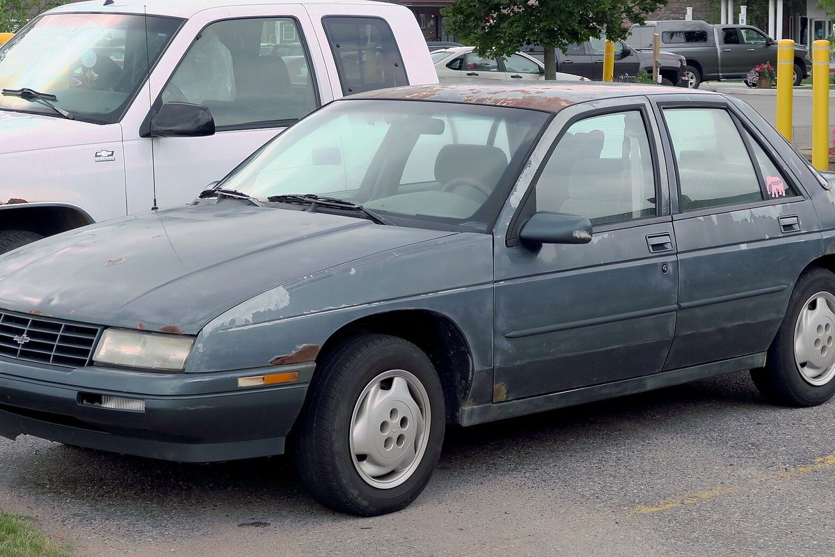 A weathered dark green-gray 1996 Chevrolet Corsica sedan with peeling paint and rust spots, parked beside a white pickup