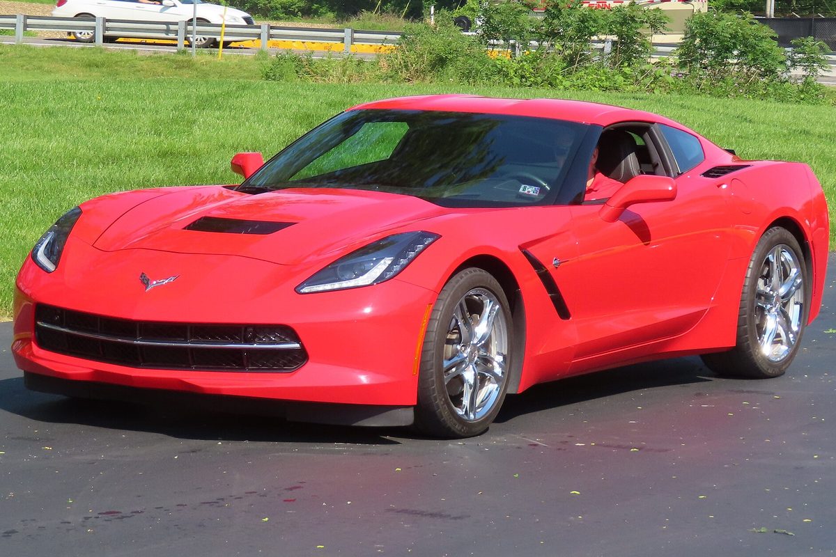 A red 2016 Chevrolet Corvette Stingray coupe with chrome wheels, parked on wet blacktop next to a mowed grass field