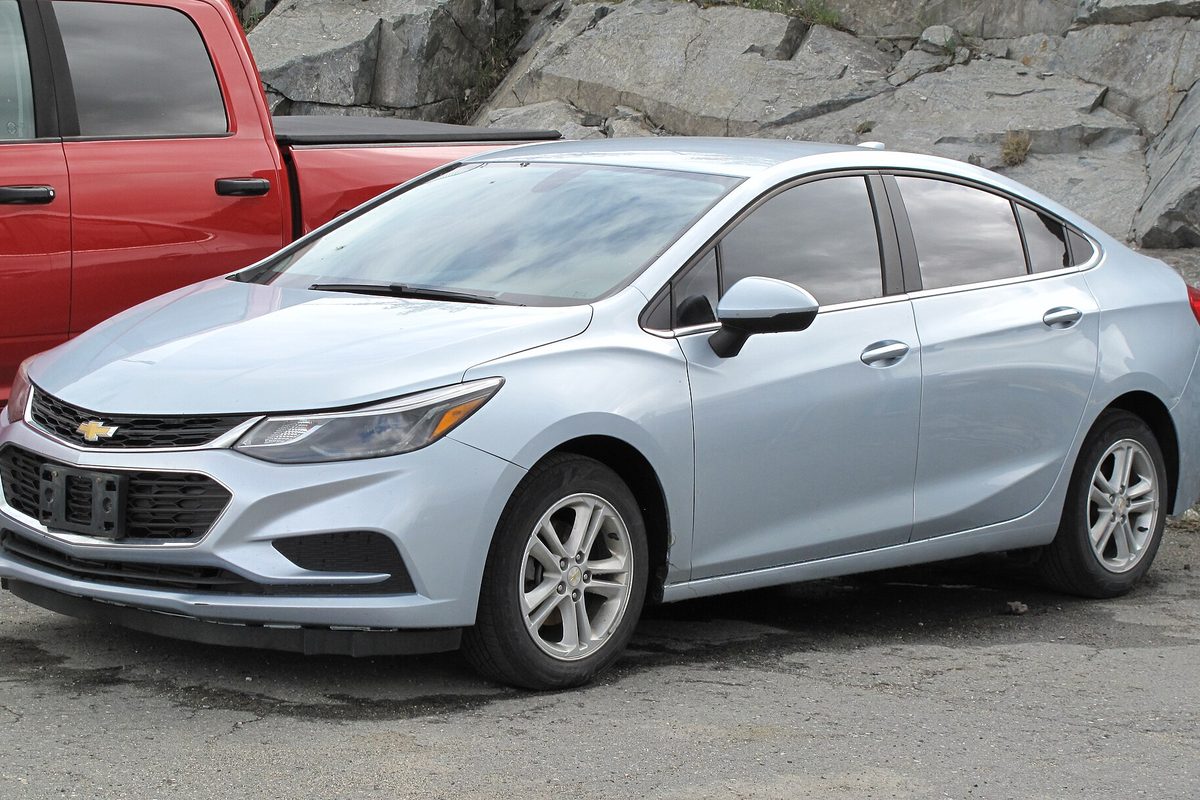 A light blue 2017 Chevrolet Cruze LT sedan parked on gravel next to a red pickup, with a gray rock cut behind it