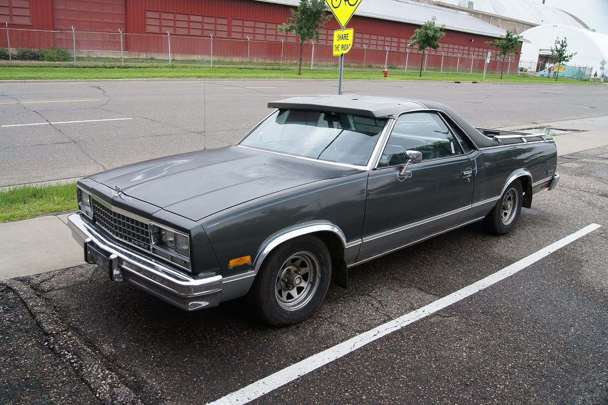 A dark gray 1984 Chevrolet El Camino diesel with chrome trim and a covered bed, parked at a curb beside a road