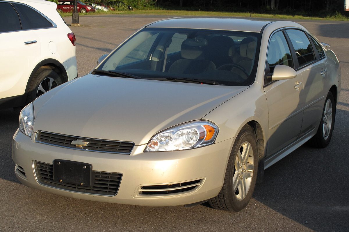 A gold 2011 Chevrolet Impala LT sedan with an empty front plate bracket, parked on an asphalt lot in low evening sun