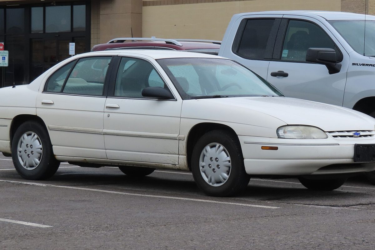 A white 2001 Chevrolet Lumina sedan with plastic wheel covers, parked in a store lot in front of a white Silverado pickup