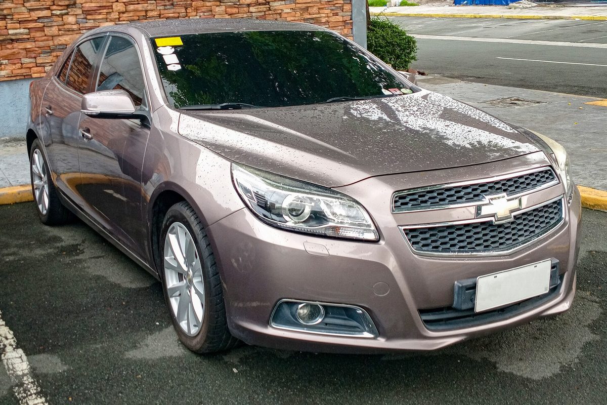 A rain-spotted bronze eighth-generation Chevrolet Malibu sedan parked in an open lot beside a stone wall