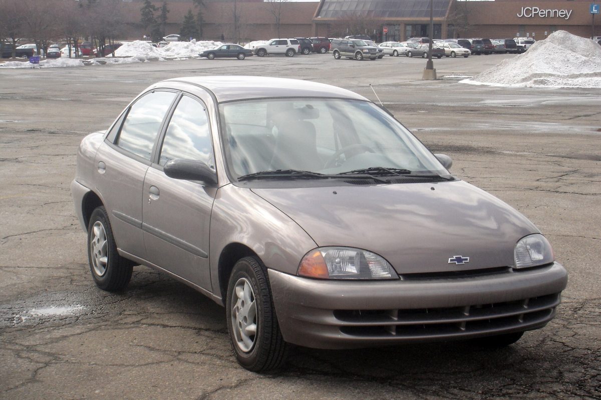 A light brown 1998 Chevrolet Metro four-door sedan in a cracked, slushy parking lot, with snow piles and a department store behind it