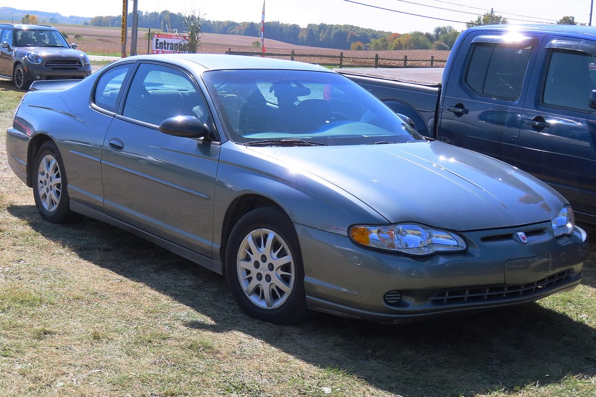 A medium gray 2004 Chevrolet Monte Carlo LS coupe parked on grass beside a blue pickup, with farm fields and trees behind