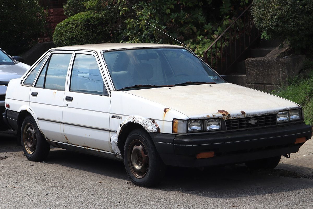 A white 1987 Chevrolet Nova sedan with rust along the hood edge and peeling paint around the front wheel arch, parked on a street below a hedge