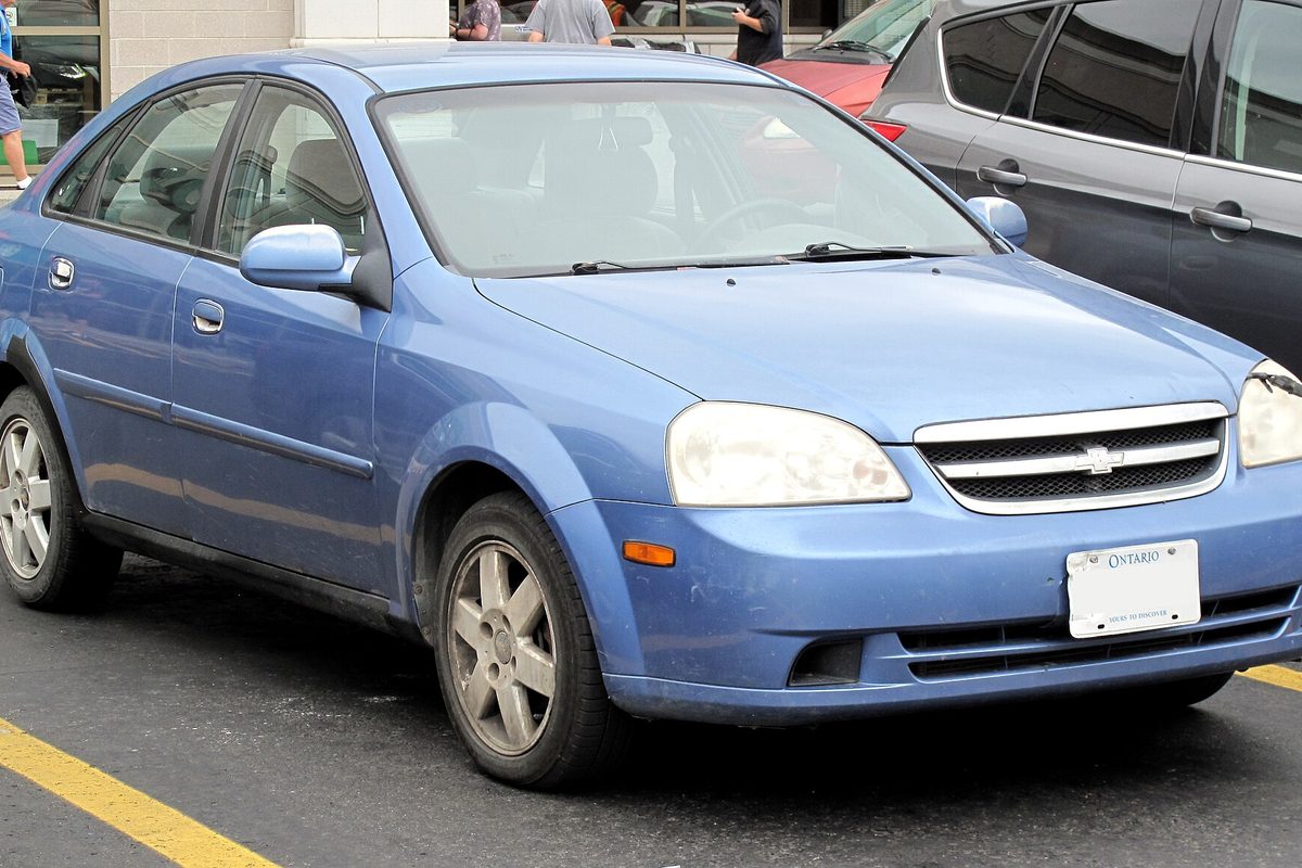 A blue 2004 Chevrolet Optra LS sedan with an Ontario plate, parked in a store lot with shoppers walking behind it