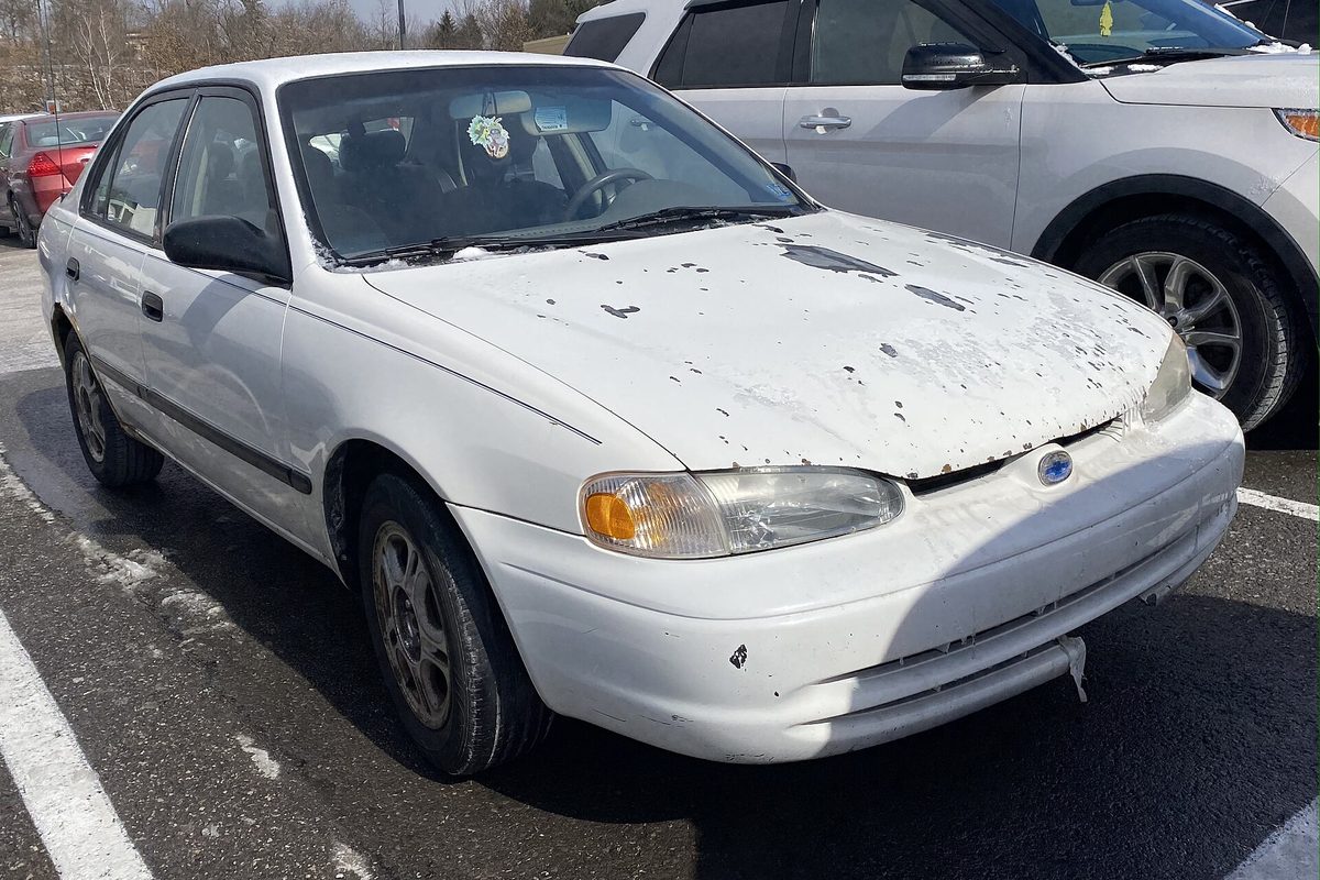 A white 1999 Chevrolet Prizm LSi sedan with paint flaking off its hood, parked in a lot next to a white SUV