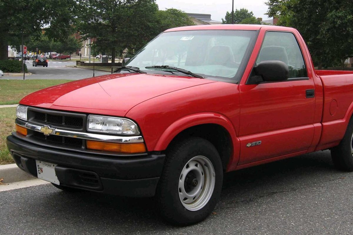 A red second-generation Chevrolet S-10 regular cab pickup with plain steel wheels, parked at a curb in front of shade trees
