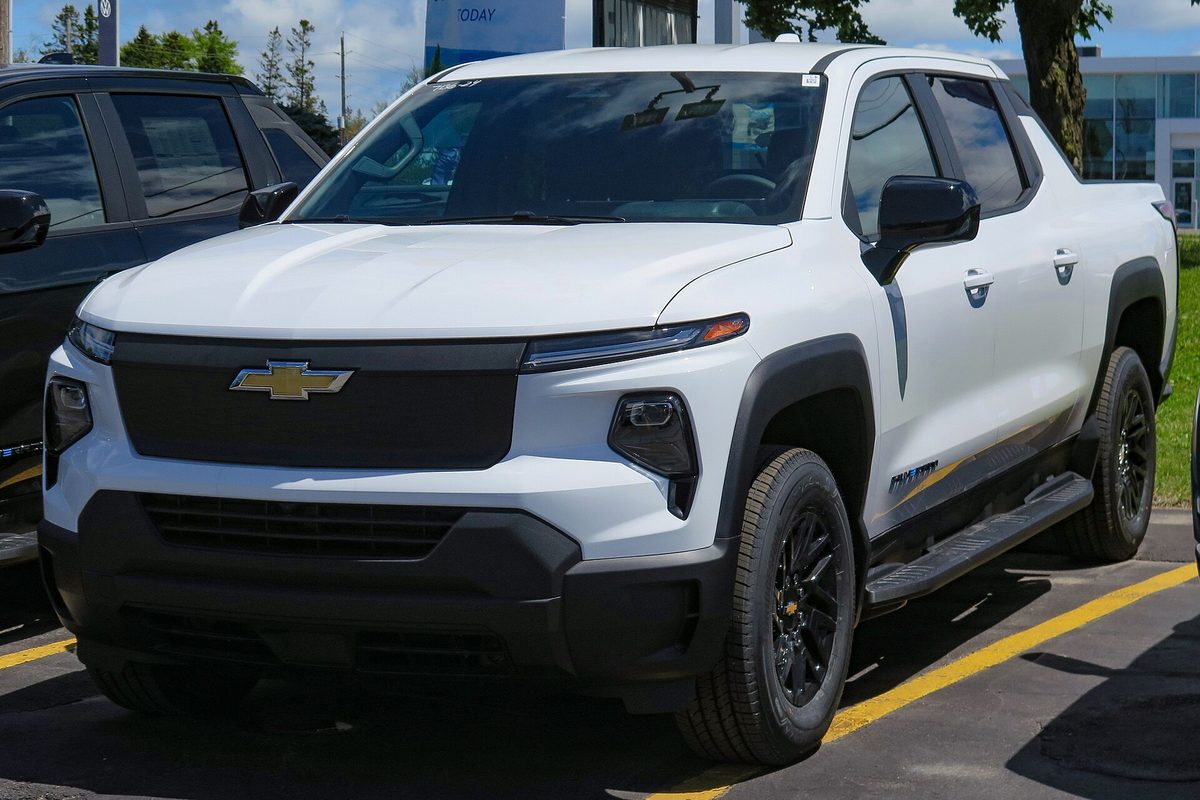A white 2024 Chevrolet Silverado EV Work Truck with black wheels and running boards, parked in a lot beside a dark SUV