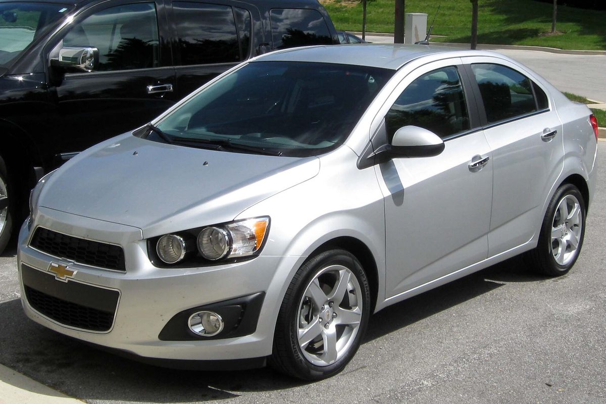 A silver 2012 Chevrolet Sonic LTZ sedan parked in a lot beside a dark SUV