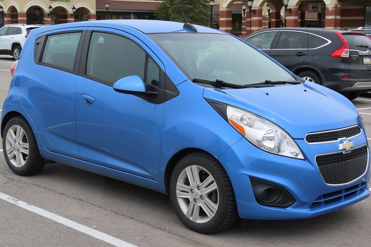 A bright blue 2015 Chevrolet Spark LS hatchback parked in a shopping center lot in front of brick storefronts