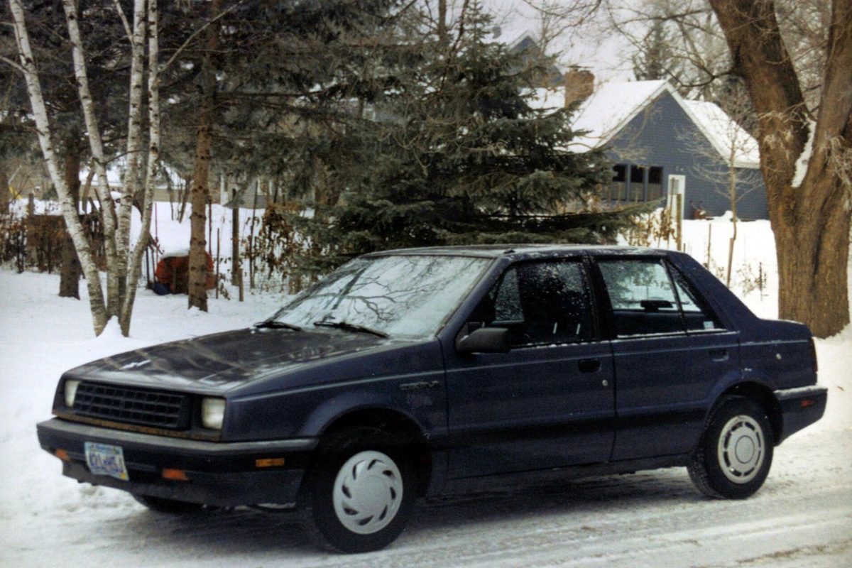A dark blue 1986 Chevrolet Spectrum sedan parked on a snowy residential street, frost on its windows, birches and pines behind it