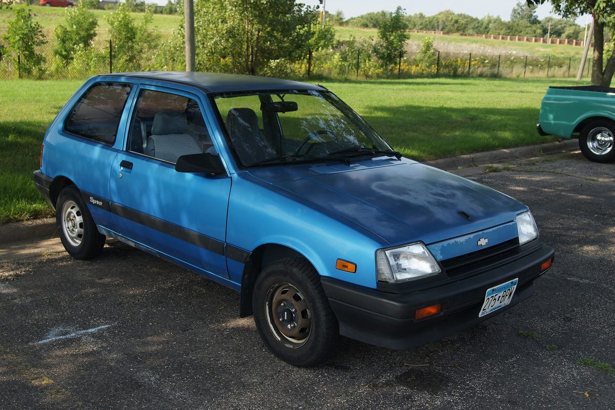 A blue 1988 Chevrolet Sprint three-door hatchback on steel wheels, parked on asphalt beside a grassy field