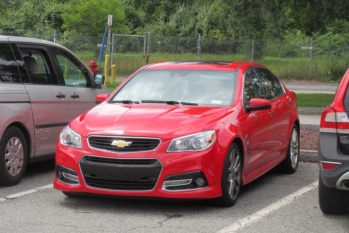 A red Chevrolet SS sedan parked in a lot beside a silver minivan, with a chain-link fence and trees behind it