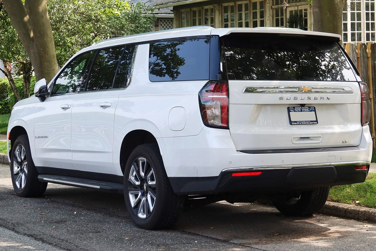 A white 2021 Chevrolet Suburban LT seen from the left rear, parked at the curb on a leafy residential street