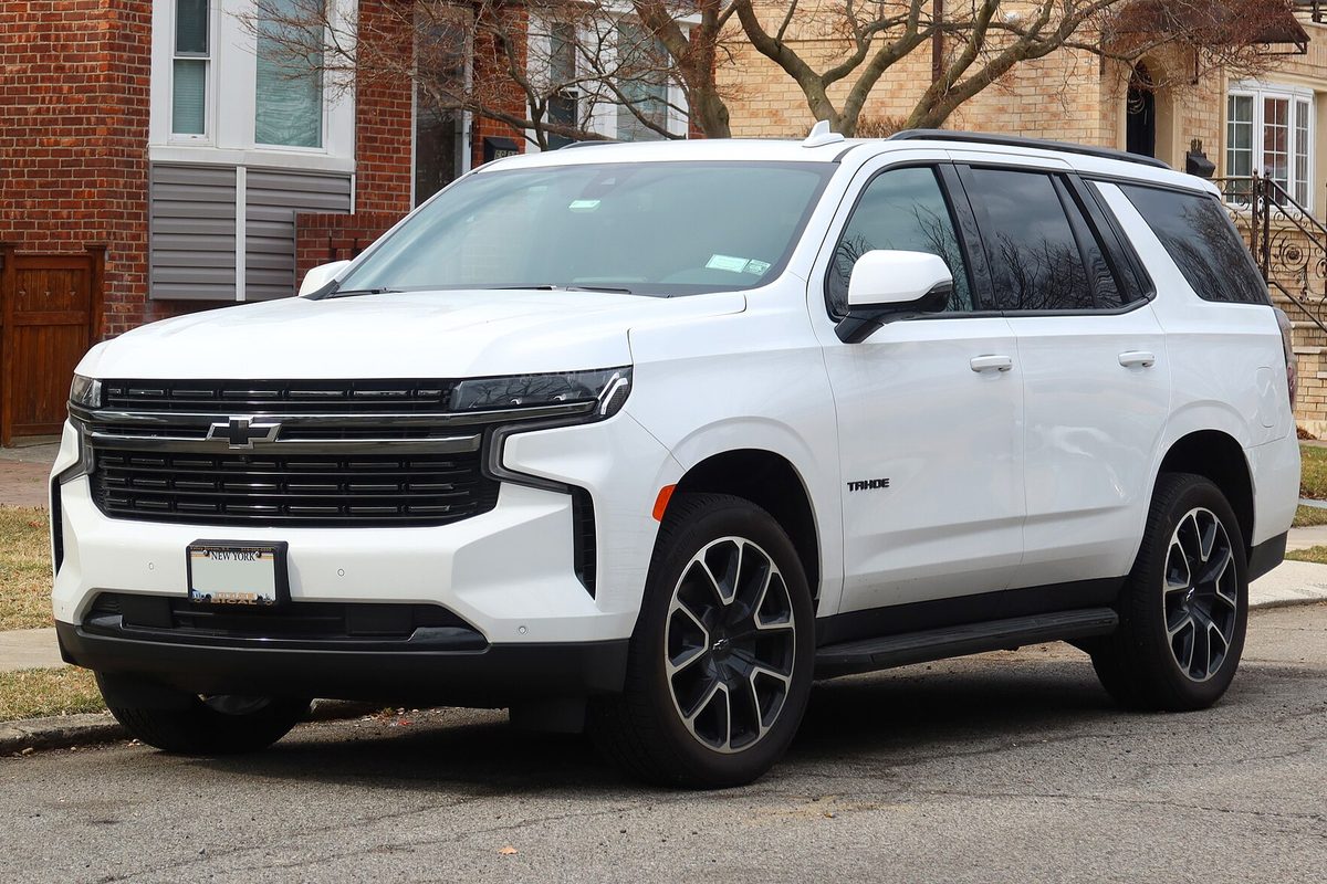 A white 2022 Chevrolet Tahoe RST with dark wheels parked on a residential street in front of brick houses and bare trees