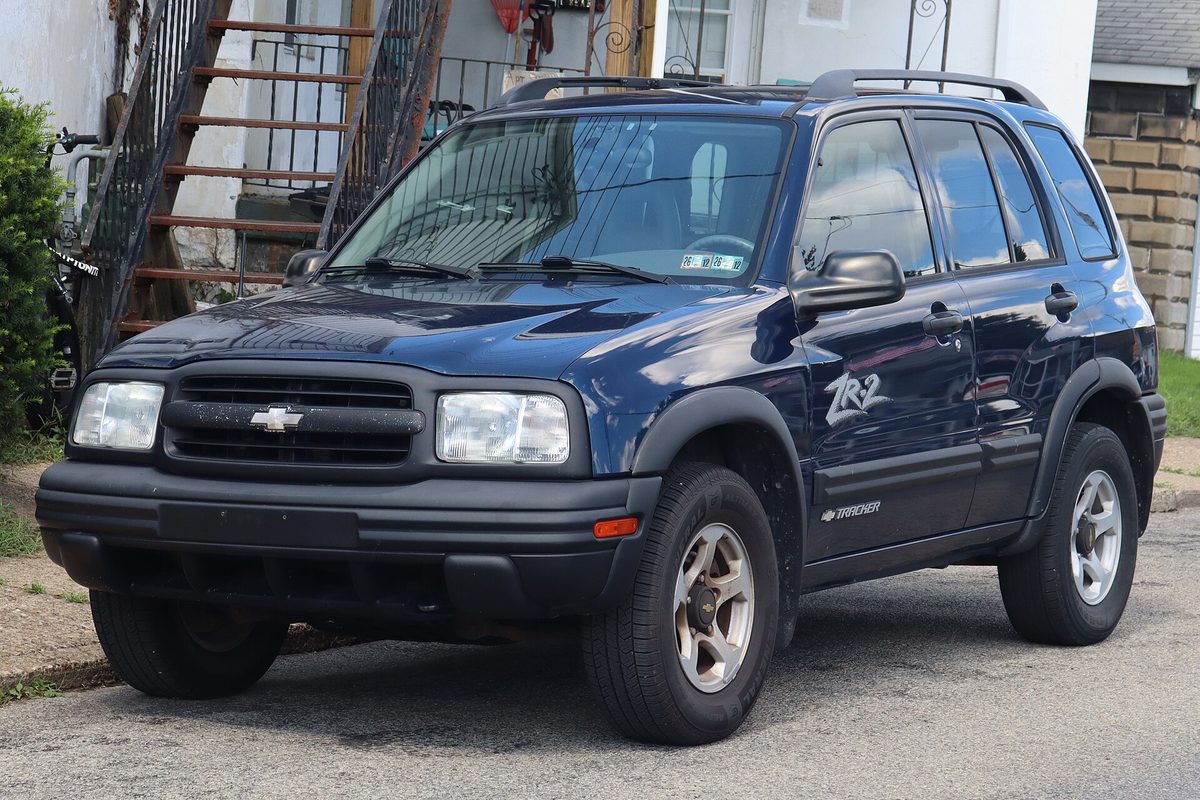 A dark blue 2002 Chevrolet Tracker ZR2 with roof rails and flared fenders, parked on a street beside wooden porch steps