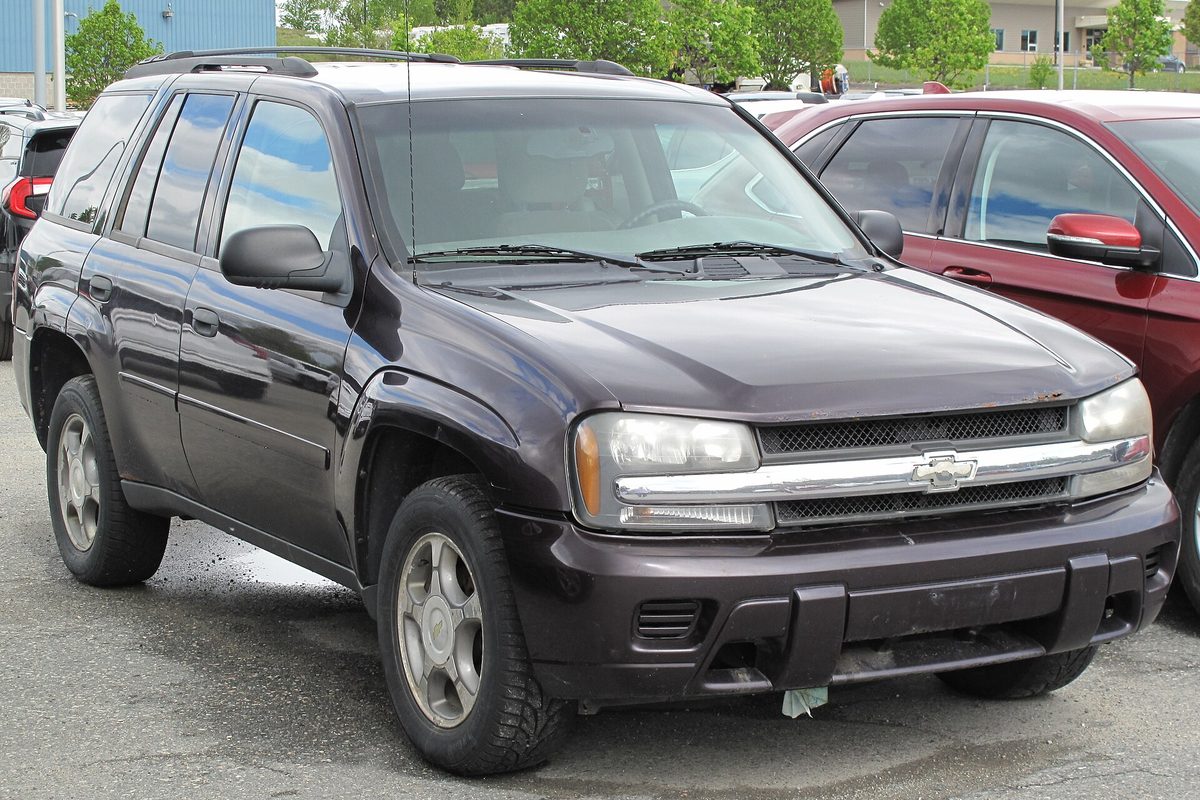 A dark cherry 2008 Chevrolet TrailBlazer LS parked on a wet lot, seen from the front right, with a red car parked behind it