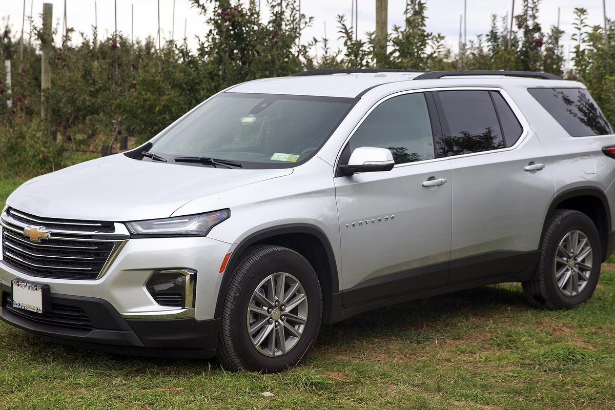 A silver 2022 Chevrolet Traverse LT, the facelifted second generation, parked on grass in front of a row of fruit trees