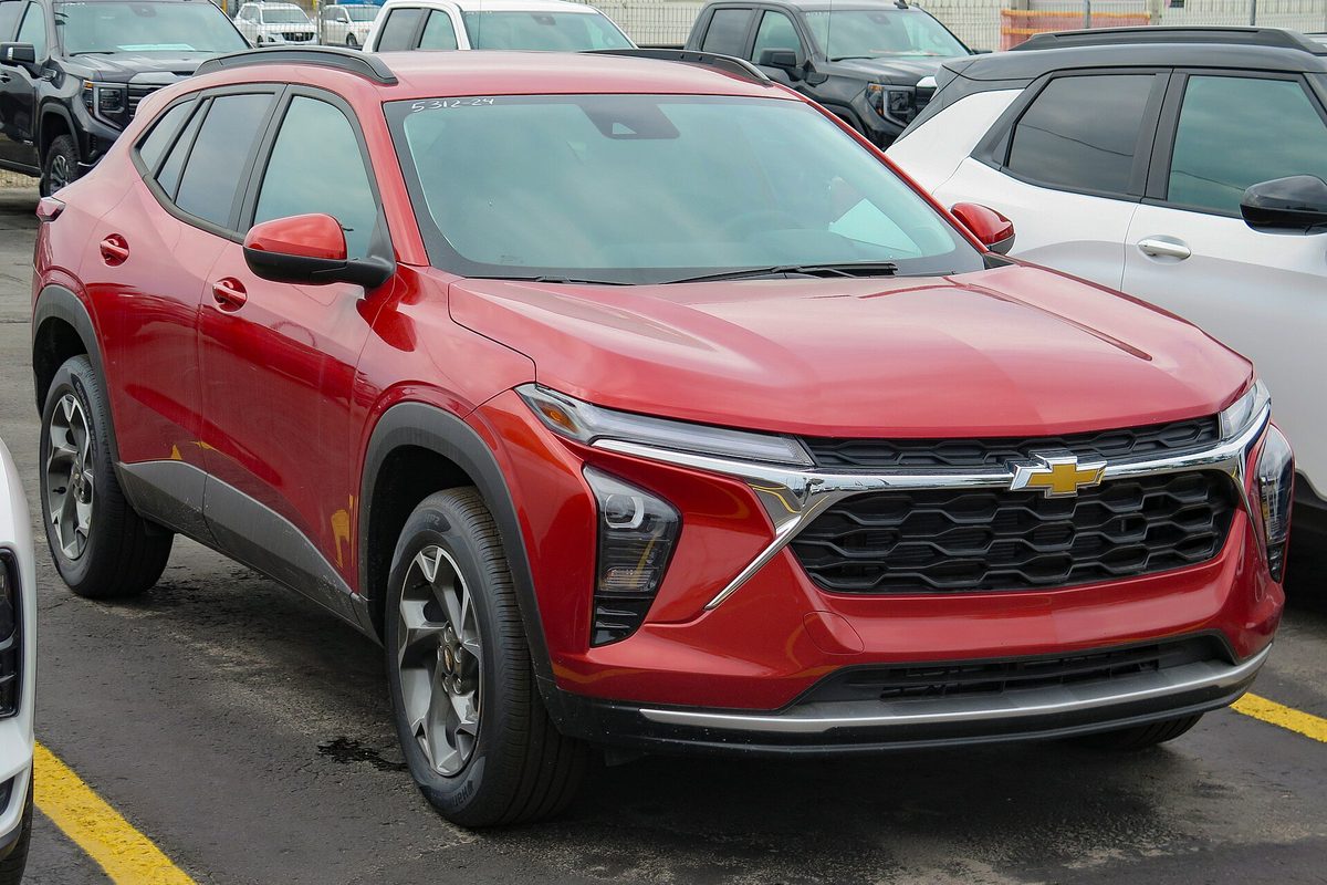 A red-orange 2024 Chevrolet Trax LT crossover parked on a dealer lot between other new SUVs