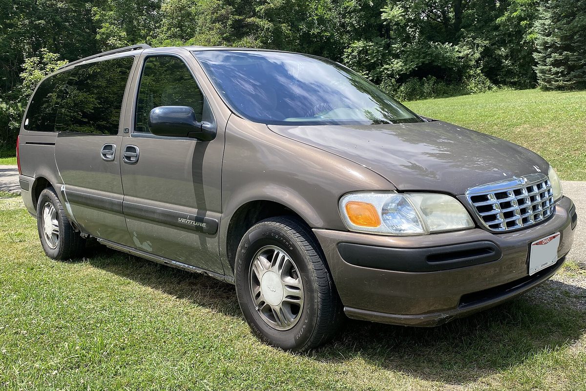 A caramel-brown 2000 Chevrolet Venture minivan with a chrome grille, parked on a sunny lawn in front of a wall of trees