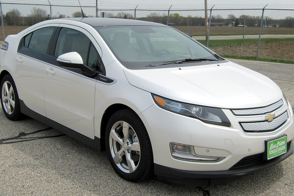 A pearl white first-generation Chevrolet Volt hatchback with silver alloy wheels, parked on cracked pavement in front of a chain-link fence