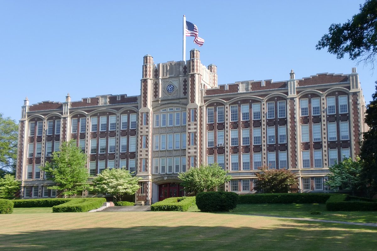 The old Chicopee High School, a long brick and stone building with a central clock tower and an American flag, behind a wide mowed lawn