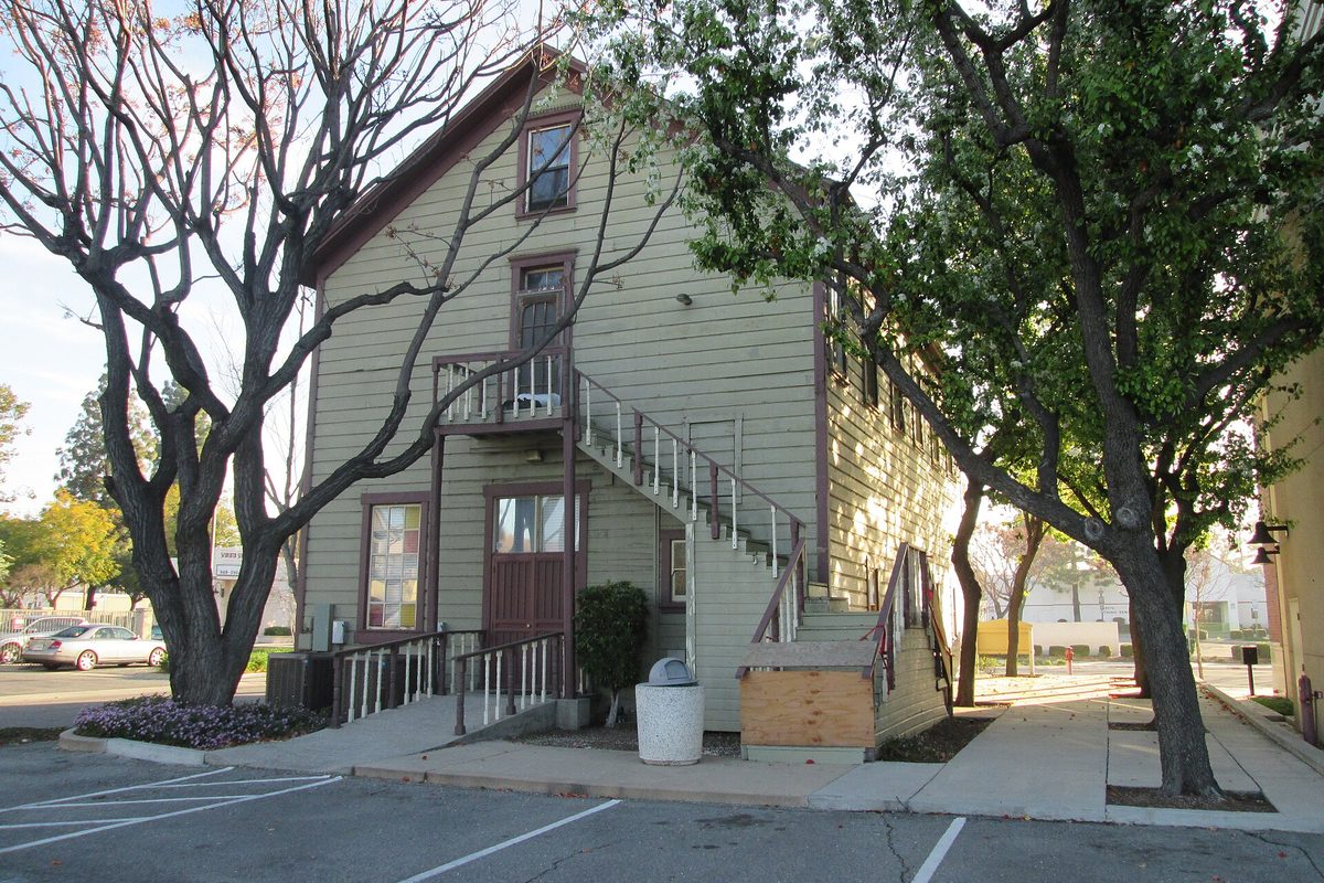 The historic two-story Moyse Building in Chino, a green wood-sided house with an outside staircase, framed by bare and leafy trees beside a parking lot