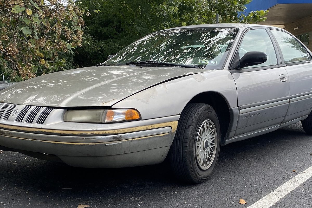 A faded light tan 1993 Chrysler Concorde sedan with a worn front bumper, parked in a lot under trees
