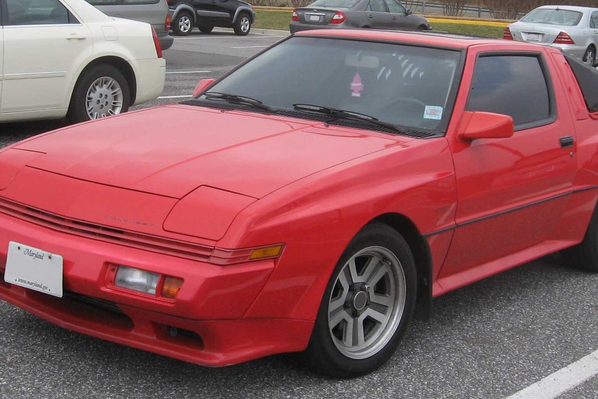 A red Chrysler Conquest hatchback coupe with flared fenders and a hood bulge, parked in a shopping-center lot