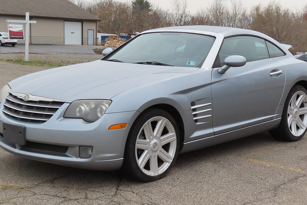 A pale blue-silver Chrysler Crossfire coupe with seven-spoke wheels parked on a cracked asphalt lot