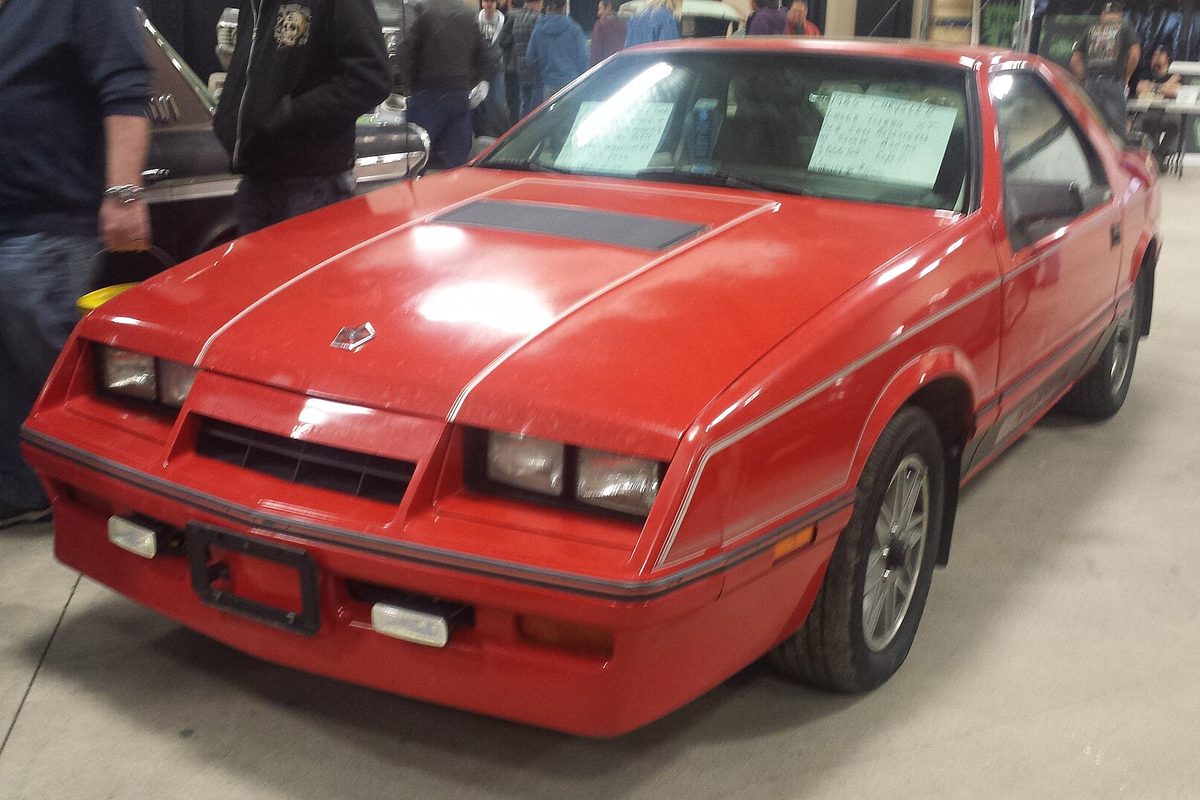 A red 1985 Chrysler Laser Turbo XT hatchback with silver pinstripes, parked indoors at a car show with for-sale notes on the windshield