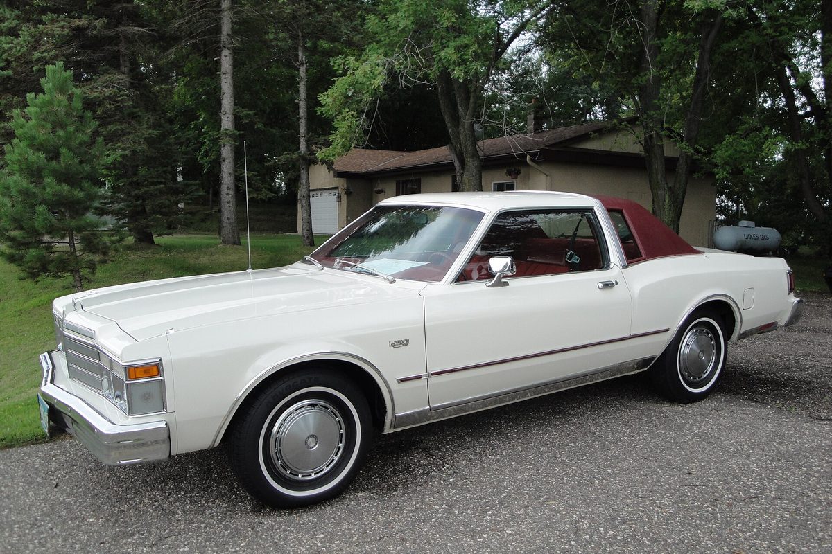A white 1979 Chrysler LeBaron coupe with a burgundy landau roof and whitewall tires on a gravel driveway