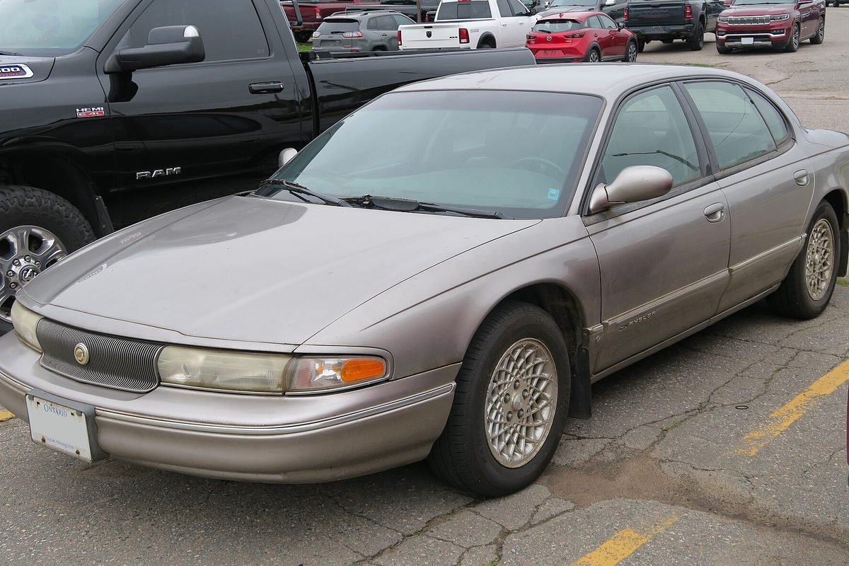 A platinum 1995 Chrysler LHS sedan with lace-style alloy wheels parked beside a black pickup on cracked asphalt
